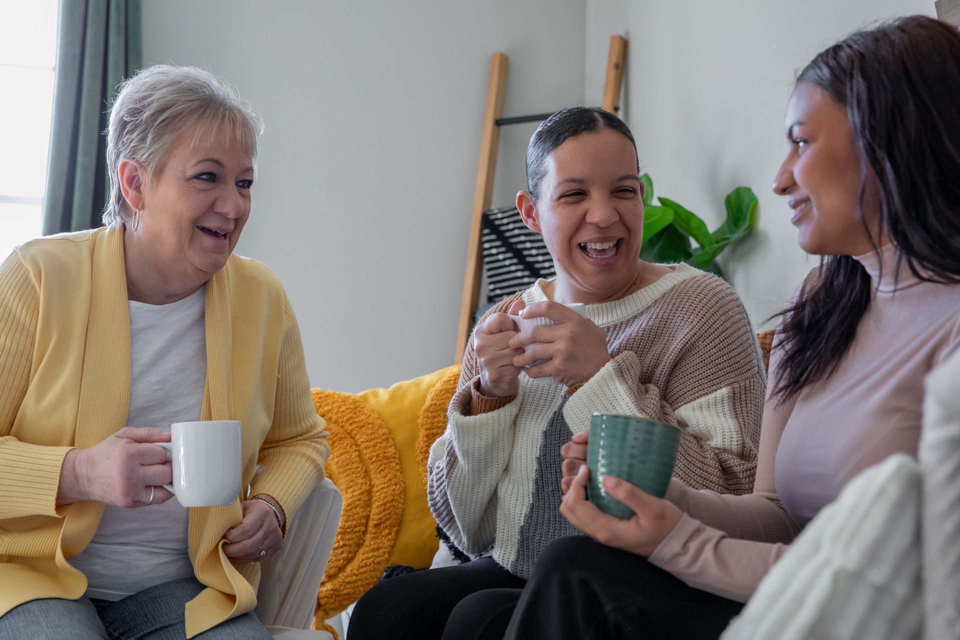 Three women, seated on a cozy couch, enjoy coffee and laugh together.